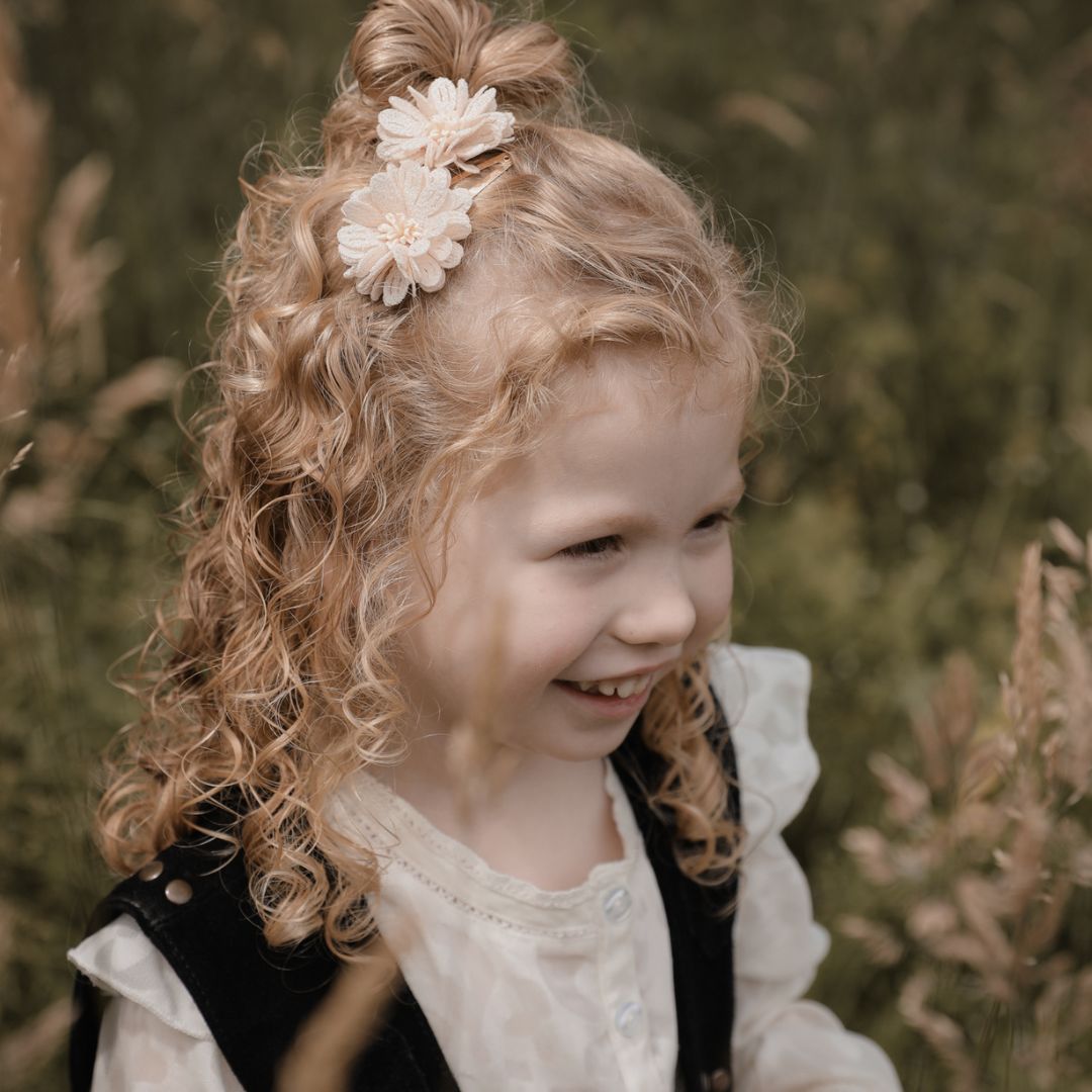 Kleuter meisje met een vrolijk krulletjes kapsel met daarin 2  goudekleurige haarspeldjes met beige bloemetjes.
